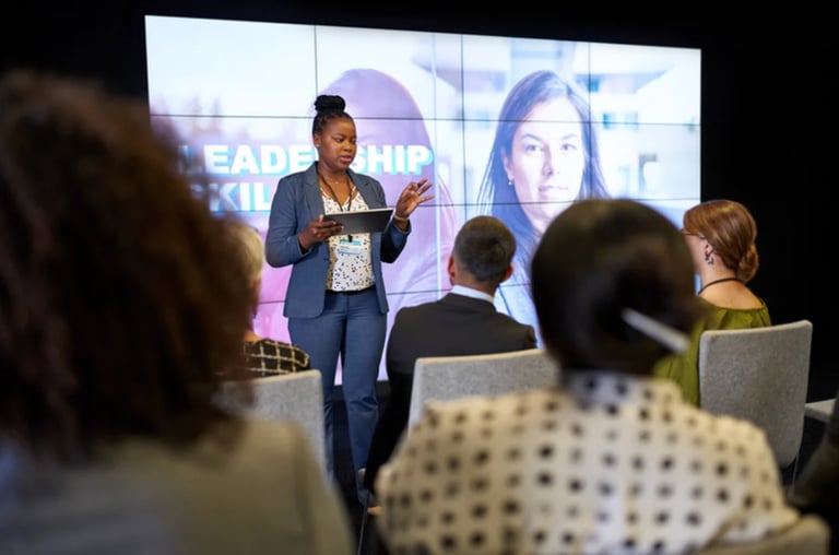 Professional woman speaking at a leadership conference, presenting to seated audience members in front of a large display screen
